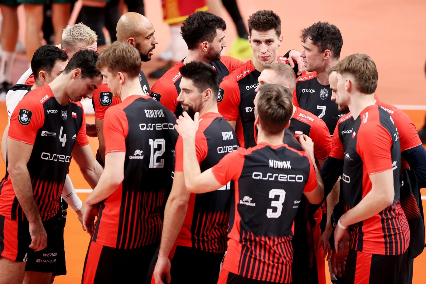 Grupa siatkarzy drużyny Asseco Resovia w czerwono-czarnych strojach zebrana w okręgu na parkiecie, prowadzą dyskusję podczas meczu siatkarskiego.