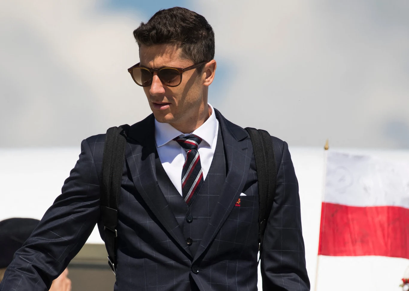 Robert Lewandowski - piłkarz FC Barcelona i reprezentacji Polski Mężczyzna w eleganckim garniturze, z okularami przeciwsłonecznymi i plecakiem na ramionach, stoi na tle flagi Polski i jasnego nieba.