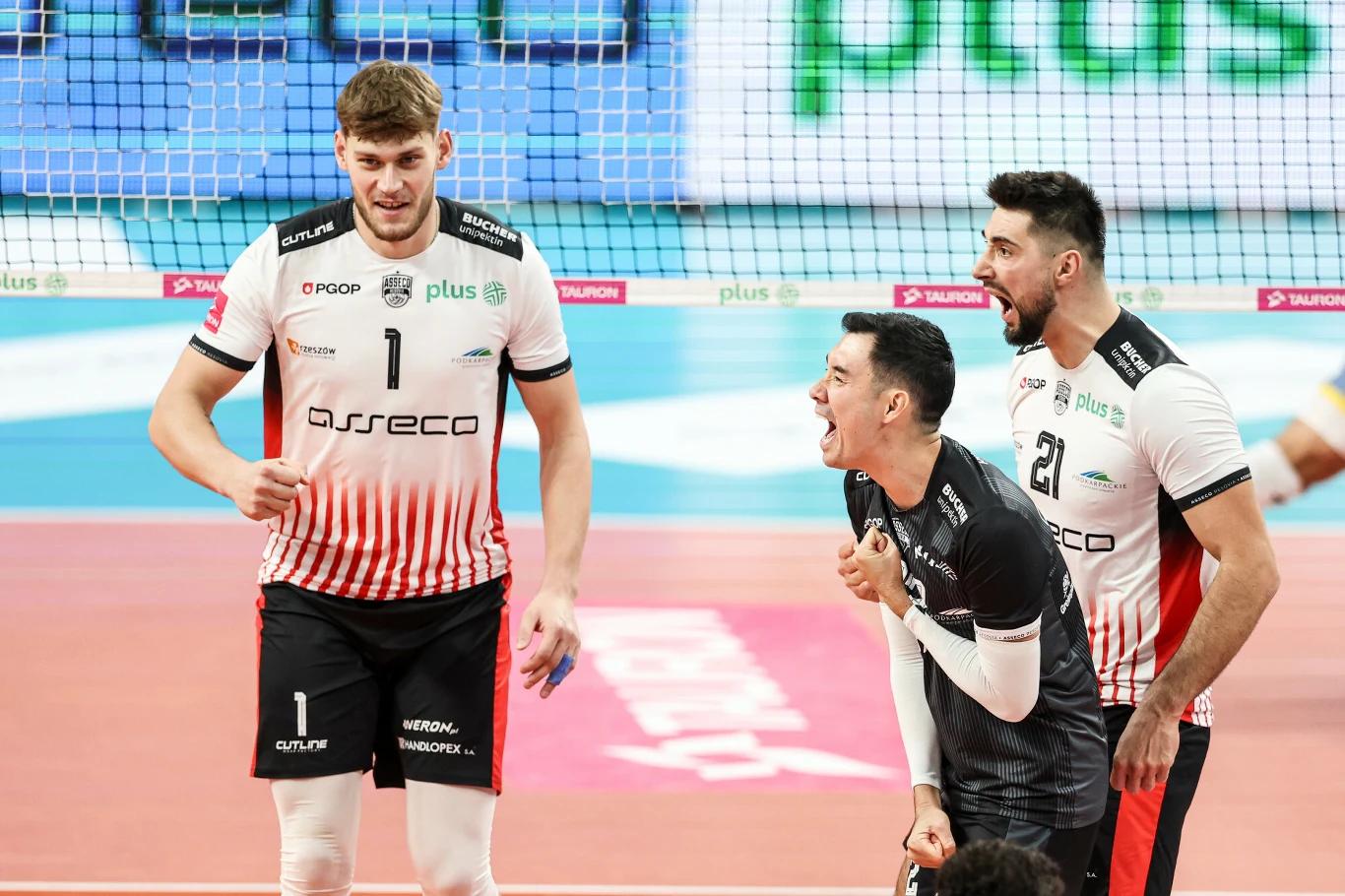 Trzech siatkarzy w strojach drużyny Asseco Resovia podczas meczu na hali sportowej, z emocjonalną reakcją jednego z zawodników oraz widoczną siatką i fragmentem boiska.