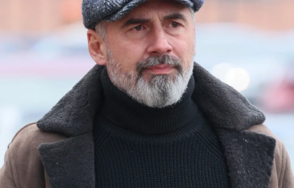 Jacek Kopczyński Mężczyzna w średnim wieku z siwą brodą i wąsami, ubrany w czarny golf oraz ciepłą, brązową kurtkę z kożuchem, na głowie ma szarą czapkę z daszkiem. Tło jest rozmyte, skupiając uwagę na osobie.