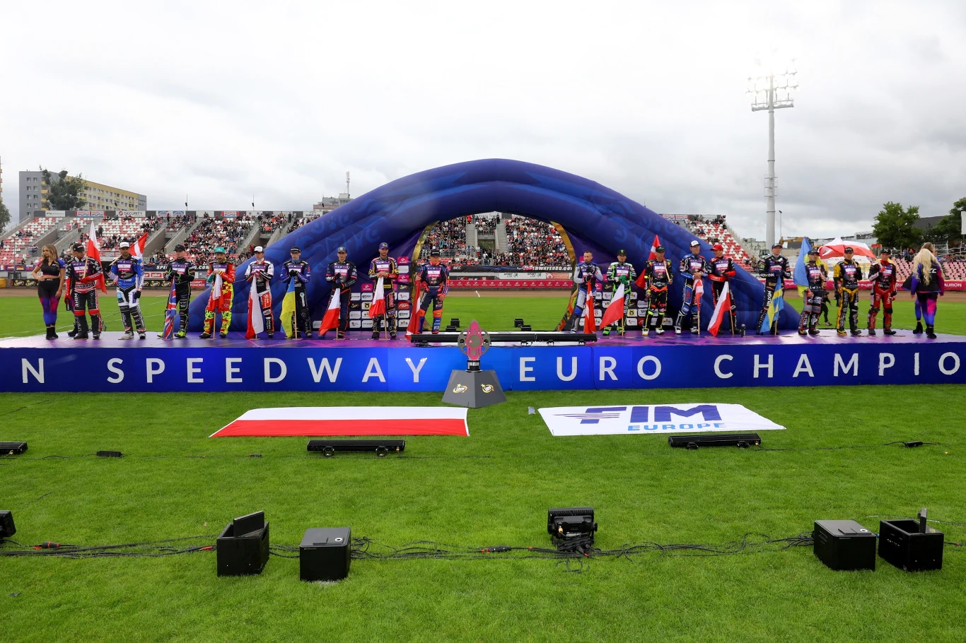 Zawodnicy żużlowi ustawieni w szeregu na podium przed rozpoczęciem zawodów, za nimi duży niebieski łuk i stadion wypełniony kibicami, na trawie widoczne flagi i logo organizatora.