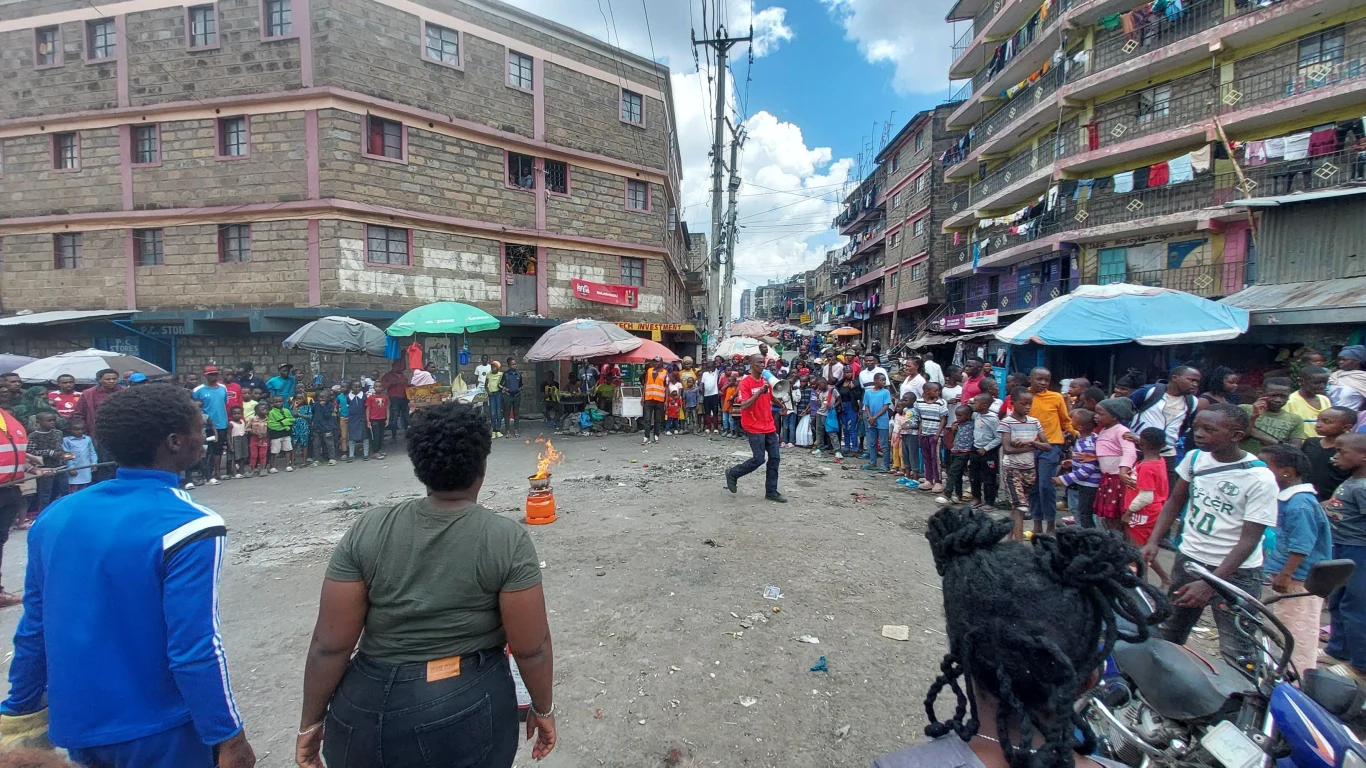 Tłum ludzi zgromadzony wokół osoby wykonującej pokaz z ogniem na środku ulicy, w otoczeniu miejskich budynków i stoisk, przy słonecznej pogodzie.