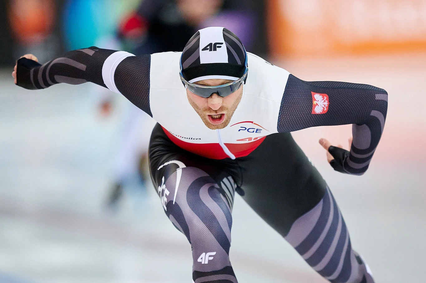 Łyżwiarz szybki w stroju reprezentacji Polski pędzi po lodowisku w dynamicznej pozycji podczas zawodów sportowych, skupiony i zdeterminowany, z widocznymi logotypami sponsorów na kombinezonie.