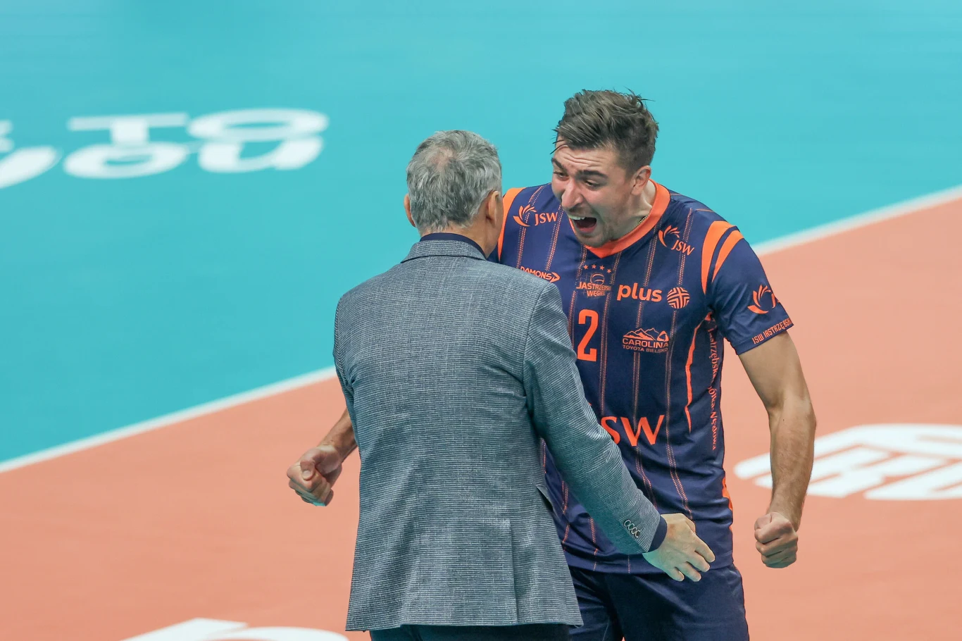 Trener Andrzej Kowal i Łukasz Kaczmarek znów mogli triumfować Zawodnik siatkarski w sportowym stroju drużyny Jastrzębski Węgiel wyraża radość w kierunku trenera na boisku, obaj w dynamicznej pozie, podkreślającej emocje meczu.