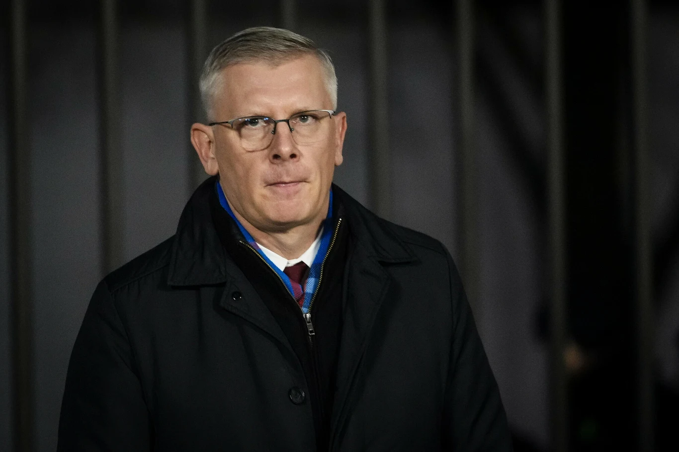 Szef BBN Sławomir Cenckiewicz Mężczyzna w okularach ubrany w elegancki płaszcz oraz krawat, patrzący prosto w stronę obiektywu, ciemne tło z pionowymi elementami metalowymi.