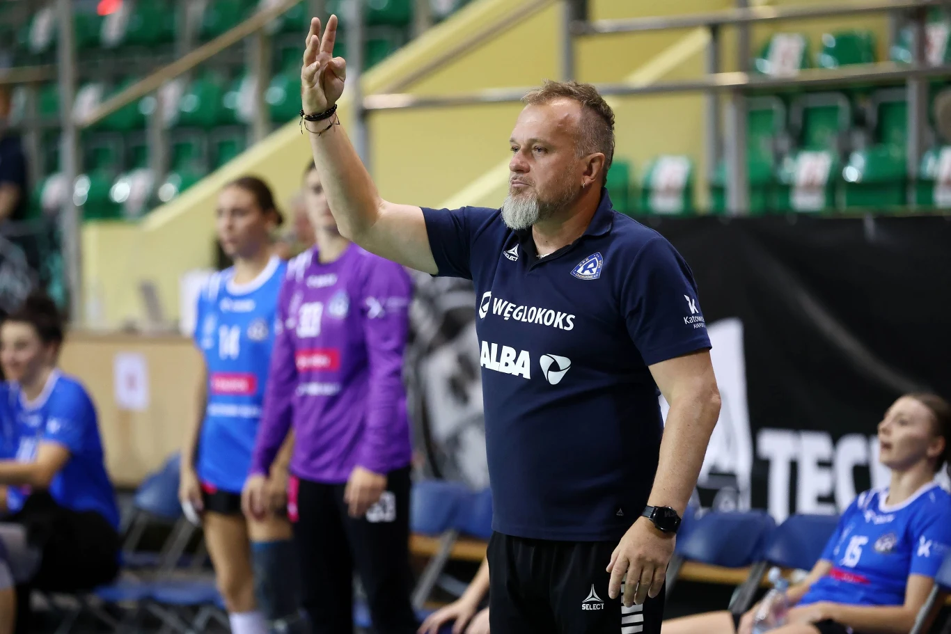 Trener piłki ręcznej w granatowym stroju wydaje polecenia na hali sportowej, unosząc dłoń z dwoma palcami w górze. W tle widoczne zawodniczki drużyny siedzące na ławce rezerwowych oraz puste zielone krzesełka trybun.