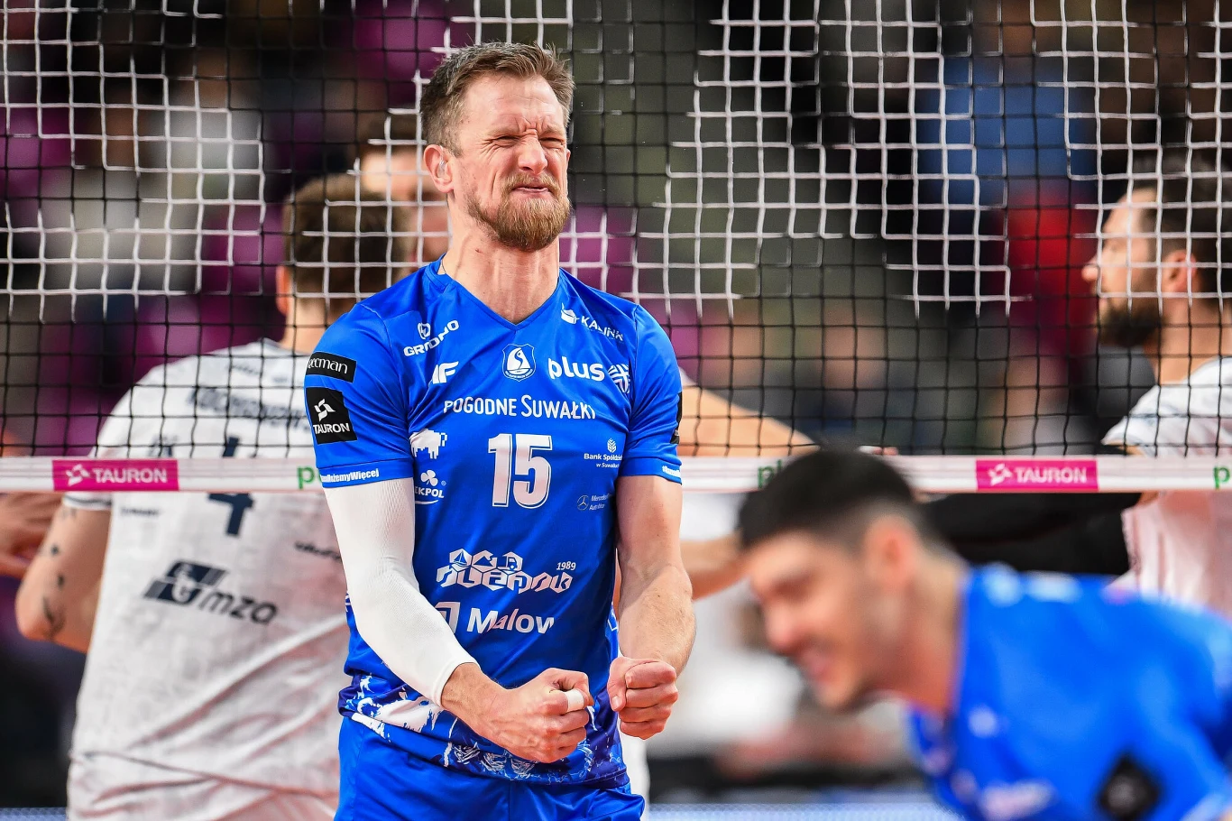 Siatkarz w niebieskim stroju drużynowym z zaciśniętymi pięściami i wyrazem intensywnych emocji na twarzy stoi przy siatce podczas meczu, wokół znajdują się inni zawodnicy.