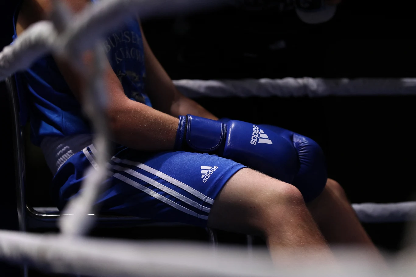 Bokser w niebieskim stroju sportowym siedzący na krześle przy linach ringu, z rękawicami bokserskimi na dłoniach, oświetlony punktowo w ciemnej scenerii.