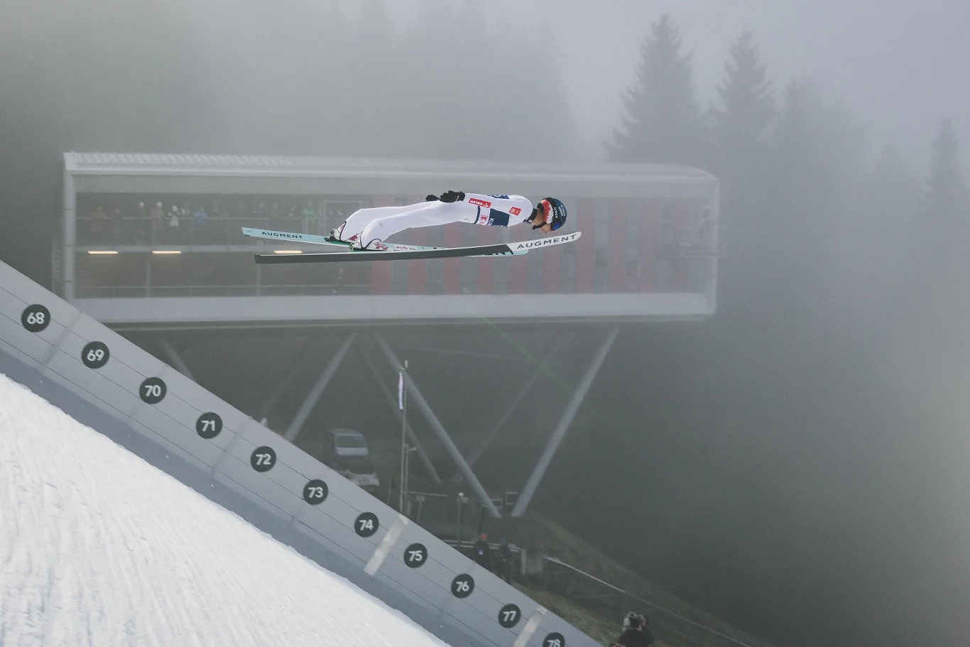 Skoczek narciarski w trakcie lotu nad skocznią, gęsta mgła, w tle nowoczesna konstrukcja wieży sędziowskiej oraz sylwetki drzew.