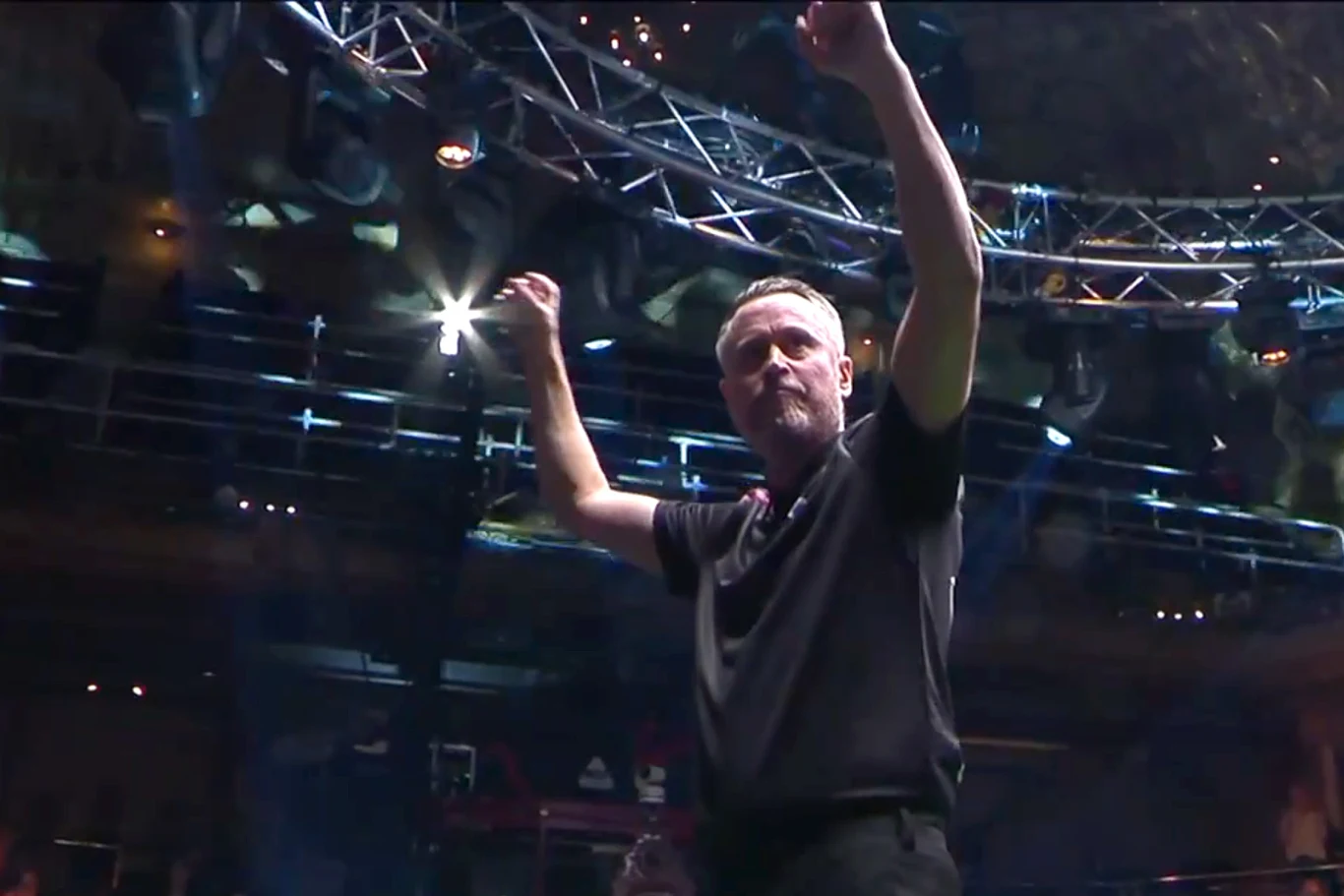 Mężczyzna w czarnej koszulce unosi ramiona w górę w geście zwycięstwa, stojąc pod metalową konstrukcją oświetleniową w hali.