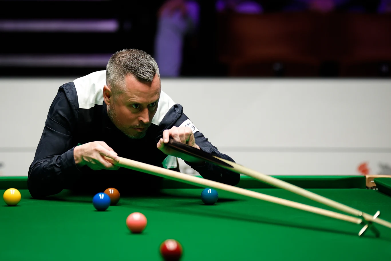 Zawodnik w skupieniu celuje kijem biliardowym do uderzenia podczas rozgrywki w snookera, na zielonym stole widoczne są rozłożone kolorowe bile.