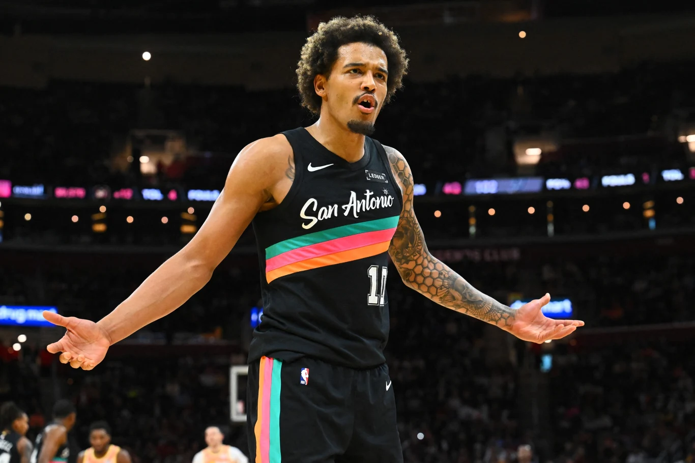 Koszykarz drużyny San Antonio Spurs w czarnym stroju z kolorowymi paskami wykonuje gest rozkładania rąk podczas meczu na hali sportowej. W tle widoczni inni zawodnicy oraz rozświetlone trybuny.