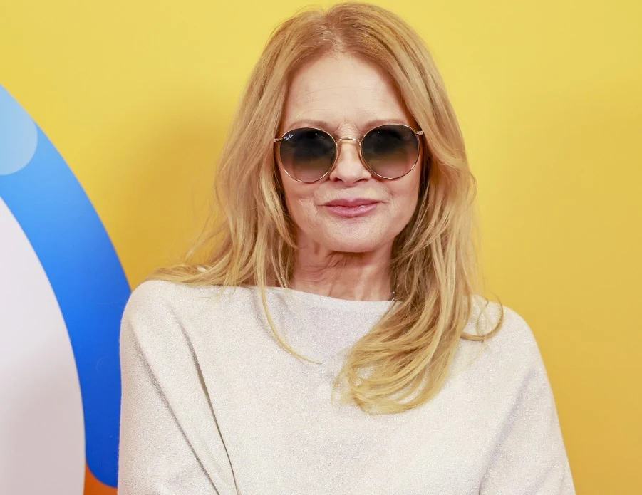 Urszula Kobieta z długimi blond włosami w okrągłych okularach przeciwsłonecznych oraz białej bluzce stoi na tle żółtej ściany z kolorowym graficznym elementem po lewej stronie.