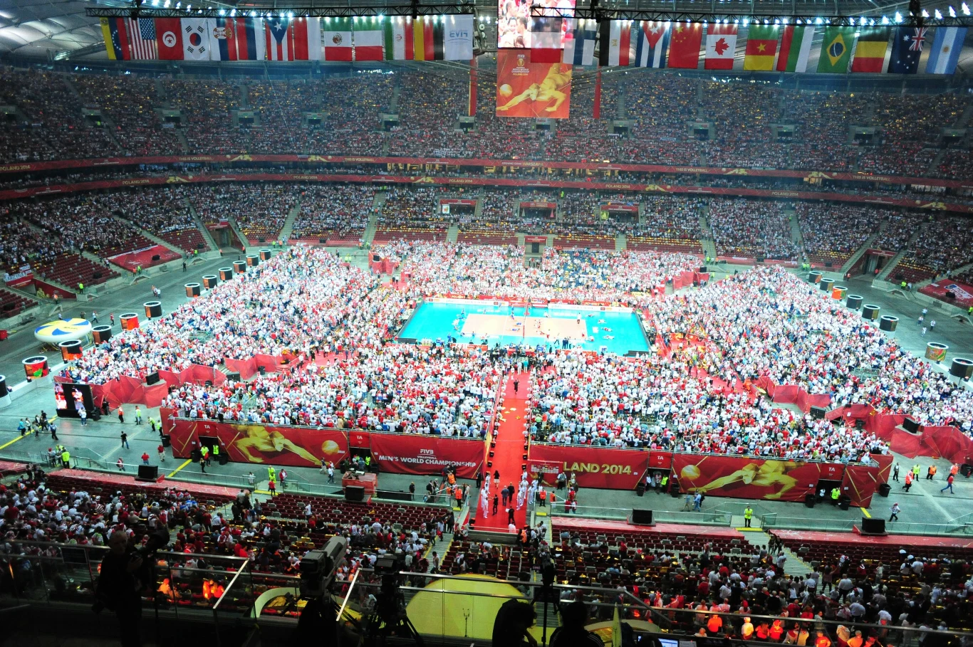 Tłum kibiców zgromadzony na stadionie wokół boiska do siatkówki, dekoracje i banery nawiązujące do mistrzostw świata, widoczne flagi państw z całego świata zawieszone nad trybunami.