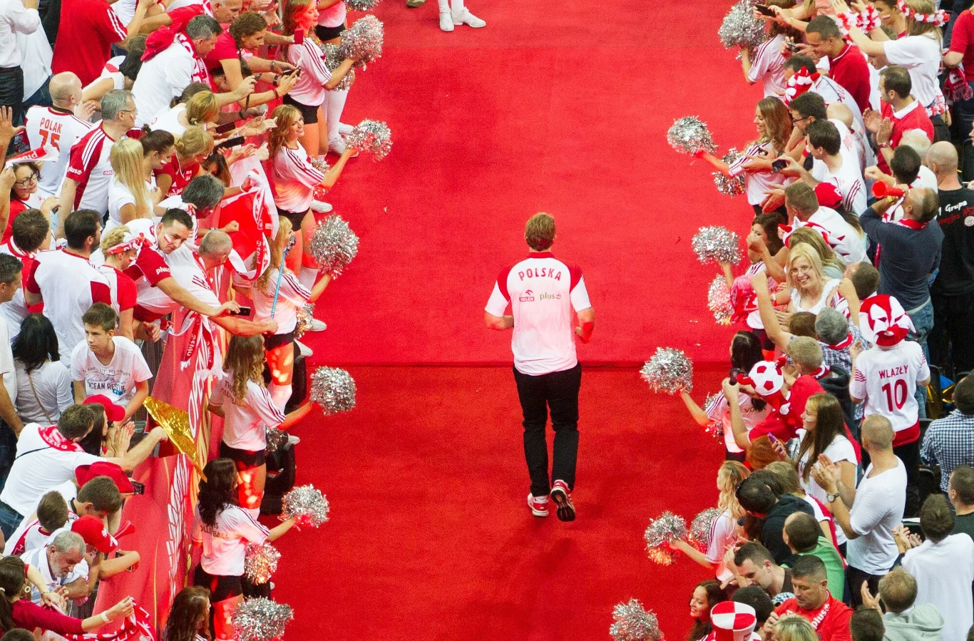 Sportowiec w białej koszulce z napisem 'Polska' idzie po czerwonym dywanie, po obu stronach kibice ubrani w biało-czerwone barwy dopingują go, machając pomponami i wyciągając ręce.