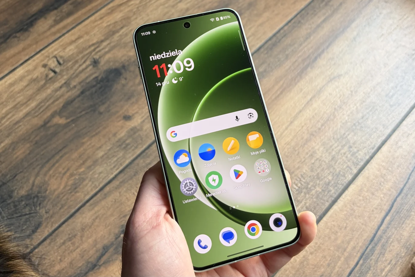 Smartfon trzymany w lewej dłoni na tle drewnianej podłogi, na ekranie widoczne ikony aplikacji, pasek wyszukiwania oraz widżet z datą i godziną.
