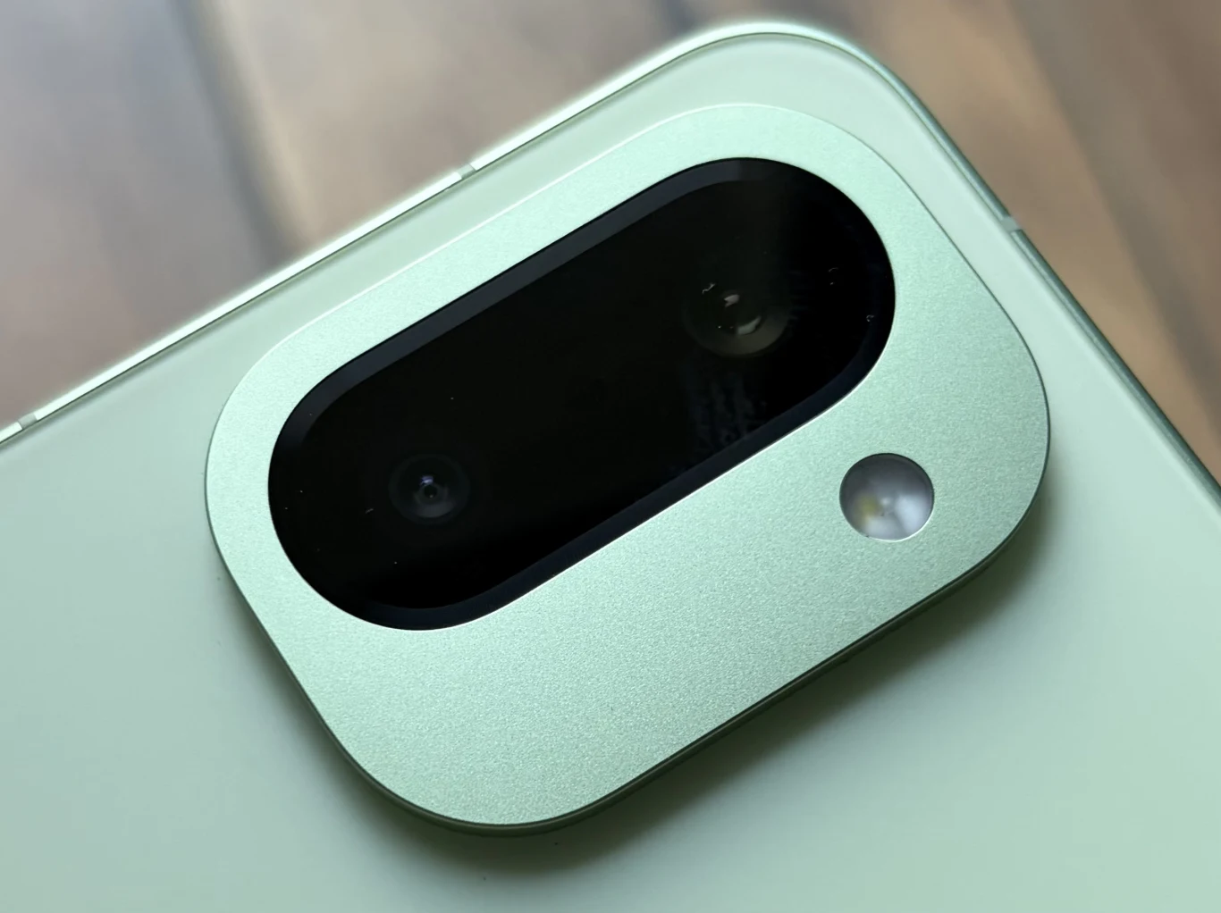 Zbliżenie na wyspę z podwójnym aparatem oraz lampą błyskową w zielonym smartfonie, aparat otoczony aluminiową ramką, tło rozmyte i neutralne.