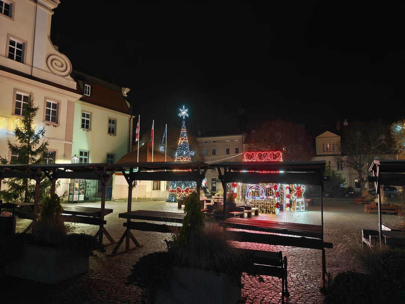 Plac miejski po zmroku udekorowany na święta Bożego Narodzenia, duża choinka z jasno świecącą gwiazdą na szczycie, iluminacje świetlne oraz budka z napisem świątecznym, otoczona przez niskie zadaszenia i ławki, budynki w tle.
