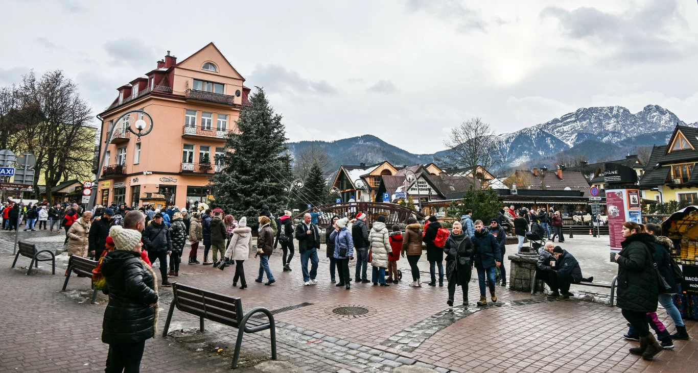 Brak odpowiednich zabezpieczeń smarfonów czy laptopów może zepsuć humor na feriach Tłum spacerujących ludzi na deptaku w mieście górskim, w tle charakterystyczna architektura budynków oraz ośnieżone szczyty górskie, przed budynkiem ustawiona choinka, atmosfera zimowa.