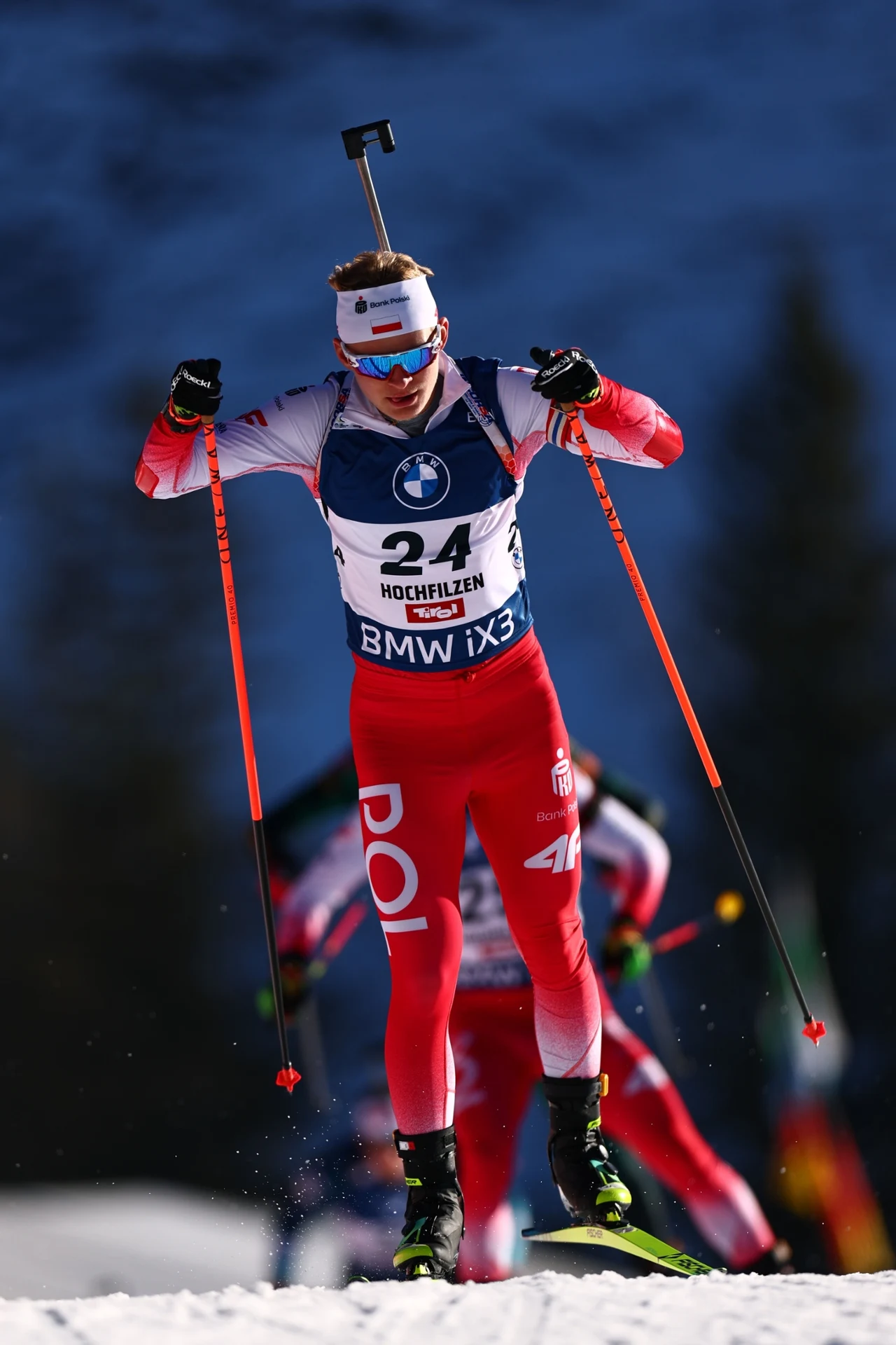 Sportowiec uprawiający biatlon w dynamicznym ruchu na trasie śnieżnej, ubrany w czerwono-biały strój z napisem POL, na głowie opaska, na plecach karabin, w tle widoczni inni zawodnicy.