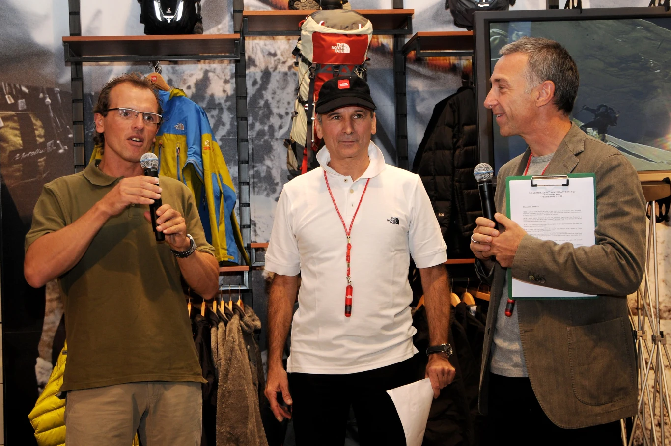 Trzech mężczyzn podczas oficjalnego spotkania lub wydarzenia, dwóch z nich trzyma mikrofony, jeden z nich ubrany na sportowo z logo marki odzieżowej na koszulce, w tle widoczne kurtki i sprzęt outdoorowy.