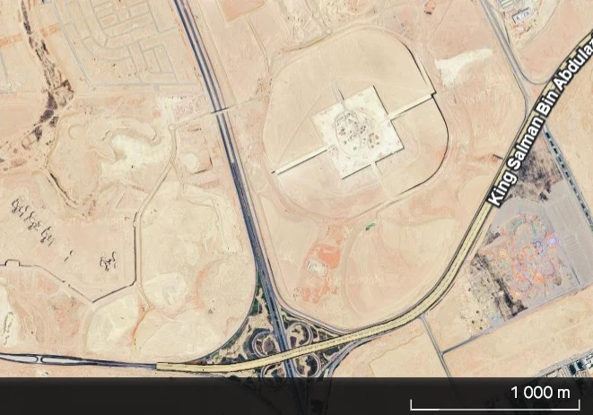 Widok satelitarny ukazujący fragment pustynnego obszaru z rozległą infrastrukturą drogową, dużym placem z geometryczną budowlą oraz wyraźnie oznaczoną ulicą King Salman Bin Abdulaziz.