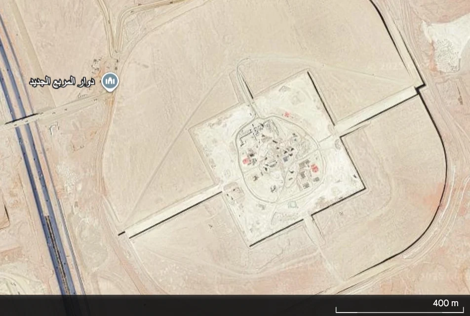 Widok satelitarny dużego, kwadratowego kompleksu otoczonego pustynią. Wewnątrz kompleksu widoczne są różne struktury i budynki, wyraźnie oddzielone od otaczającego obszaru wysokimi murami lub barierami. W pobliżu znajduje się lokalizacja oznaczona napi...