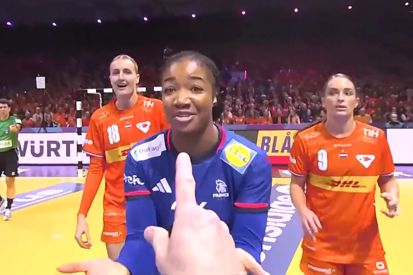 Reprezentacja Francji i Holandii stworzyły pasjonujące widowisko. O brązie decydował dodatkowy czas Sportowa rywalizacja podczas meczu piłki ręcznej kobiet, zawodniczka w niebieskim stroju stoi na pierwszym planie, dwie zawodniczki w pomarańczowych strojach obok, widoczny fragment boiska, sędzia oraz trybuny pełne kibiców w tle.