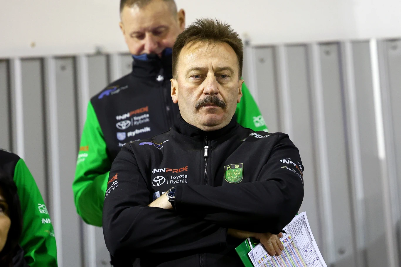 Prezes Krzysztof Mrozek Mężczyzna z wąsem w sportowej kurtce z logotypem drużyny, trzymający złożone ręce i dokumenty, stoi na tle metalowych paneli, w tle inny mężczyzna w podobnej odzieży.