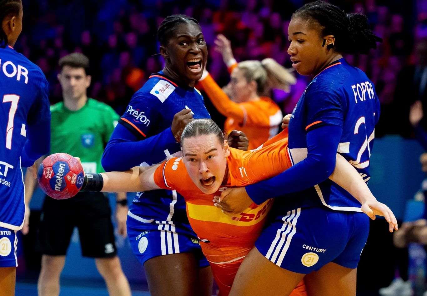 Romee Maarschalkerweerd Zawodniczki podczas dynamicznej akcji meczowej piłki ręcznej, jedna z nich w pomarańczowym stroju jest silnie blokowana przez dwie rywalki w niebieskich strojach, w tle widać sędziego oraz pozostałych uczestników meczu.