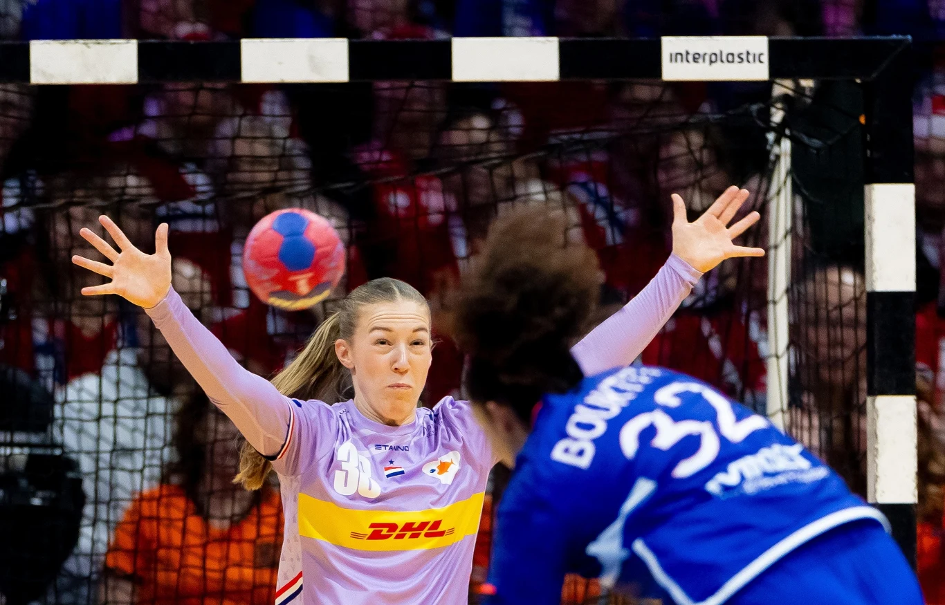 Yara Ten Holte broni, rzuca Sarah Bouktit Bramkarka w fioletowym stroju z uniesionymi rękami próbuje obronić rzut przeciwniczki w niebieskim stroju z numerem 32 podczas meczu piłki ręcznej, w tle widoczna siatka oraz kibice.