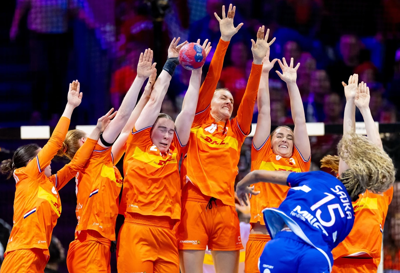 Francja - Holandia, mecz o brąż mistrzostw świata Zawodniczki drużyny w pomarańczowych strojach bronią bramki w meczu piłki ręcznej, próbując zablokować rzut zawodniczki drużyny przeciwnej ubranej na niebiesko. W tle widoczna publiczność i hala sportowa oświetlona reflektorami.