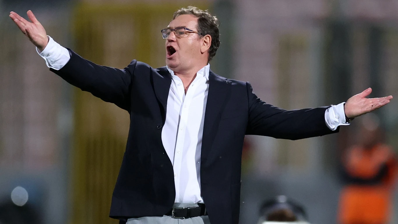 Selekcjoner Jan Urban Mężczyzna w okularach i garniturze z rozłożonymi szeroko rękami w geście wyrażającym emocje, najprawdopodobniej podczas wydarzenia sportowego, na tle rozmazanego stadionu.