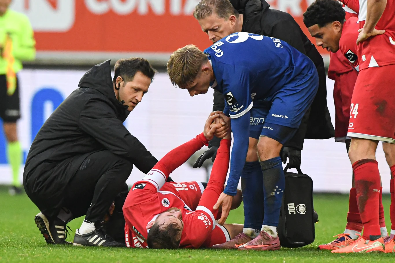 Michał Skóraś przeprasza brutalnie sfaulowanego rywala Piłkarz leży na murawie i trzyma się za ramię w wyniku kontuzji, obok klęka ratownik medyczny oraz inny zawodnik, który podaje rękę. Dwóch kolejnych piłkarzy z boku obserwuje sytuację, wszyscy skupieni są na pomocy koledze. Ubrania sportowe i sprzęt me...