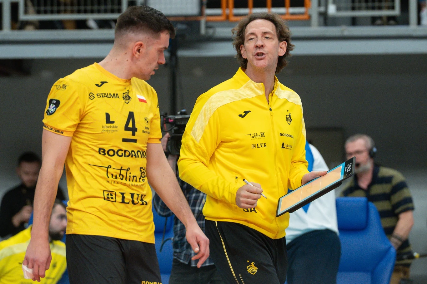 Trener w żółtej kurtce rozmawia z siatkarzem ubranym w żółty strój z numerem 4, obaj znajdują się na hali sportowej podczas meczu lub treningu, w tle widoczni są inni ludzie siedzący na niebieskich krzesłach.