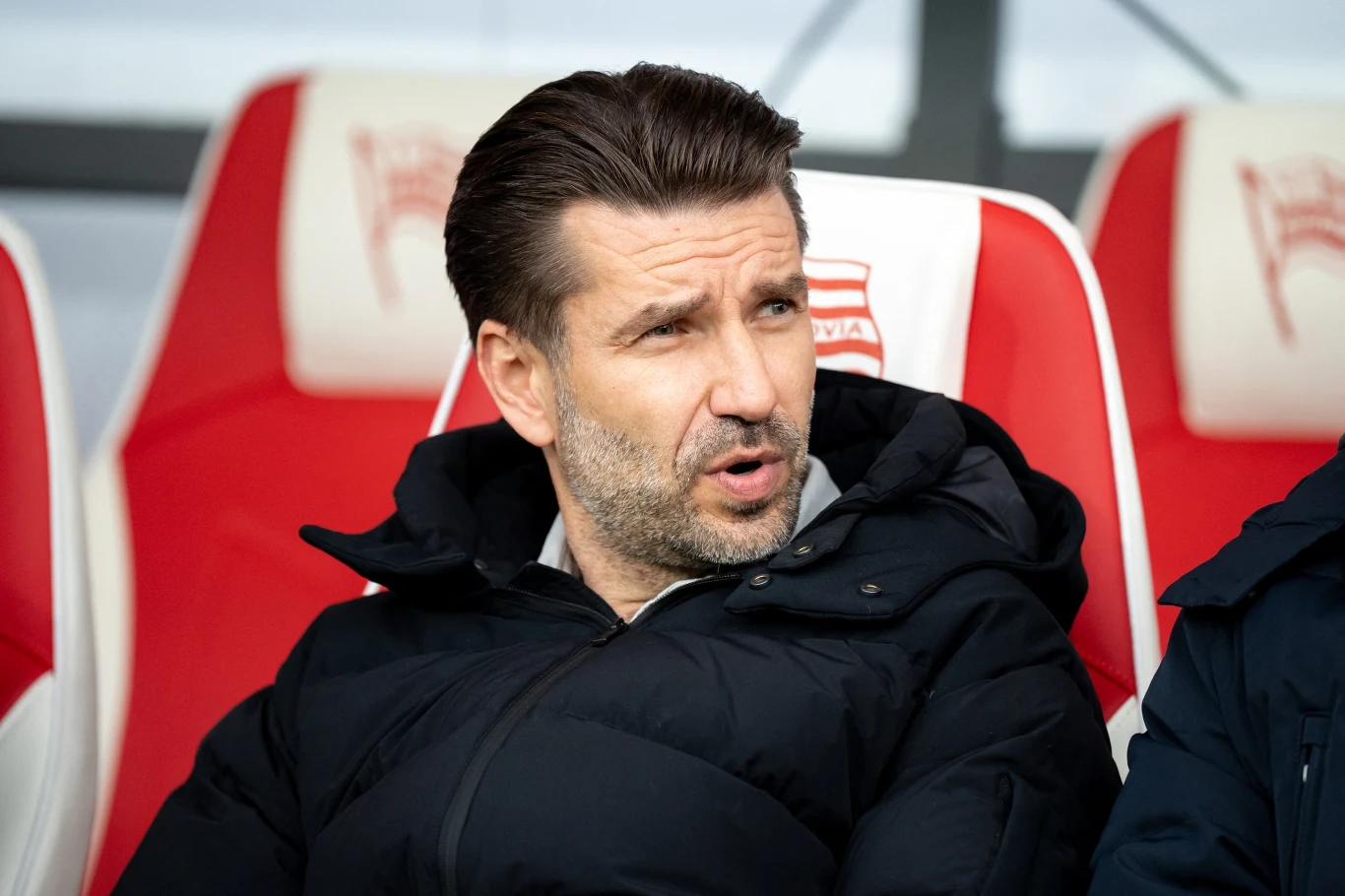 Mężczyzna z ciemnymi włosami w czarnej kurtce siedzi na stadionowej ławce trenerskiej z białym i czerwonym oparciem na tle stadionowych siedzisk.