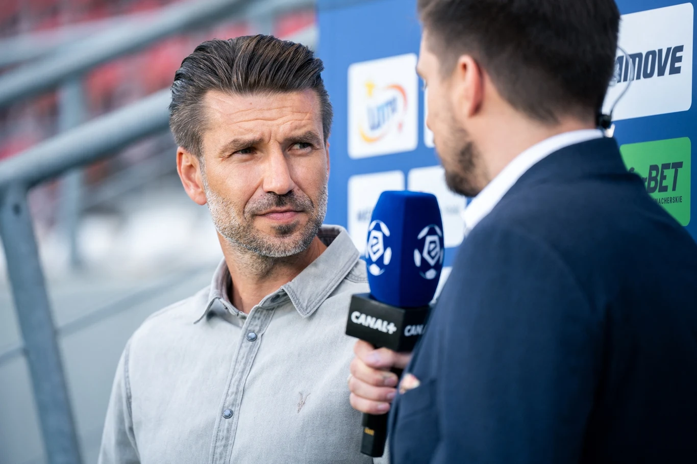 Dwie osoby prowadzą rozmowę na żywo przed kamerą, jedna z nich trzyma mikrofon z logo Canal+, w tle widoczne są barwy i logo sponsorów.