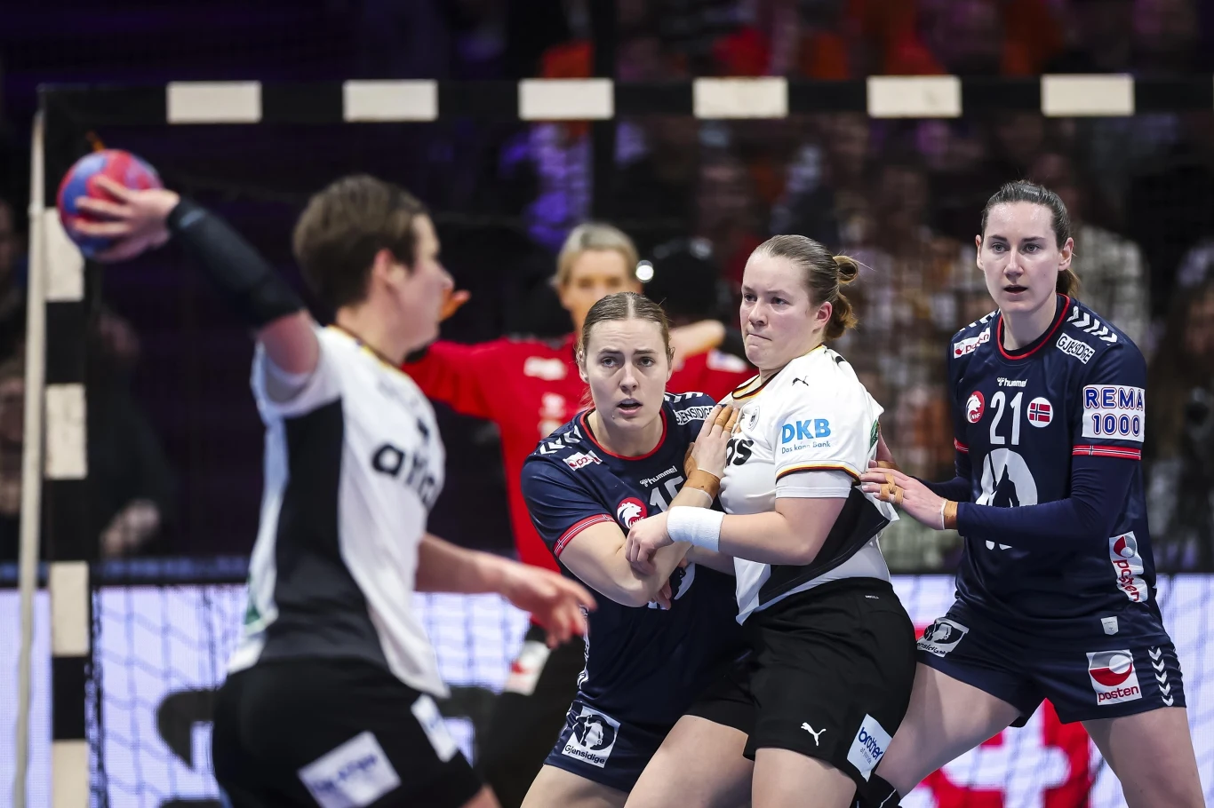 Intensywna walka podczas meczu piłki ręcznej kobiet, dwie zawodniczki w ciemnych strojach próbują powstrzymać przeciwniczkę w białej koszulce, która trzyma piłkę. W tle widać bramkę oraz skupione twarze uczestniczek.