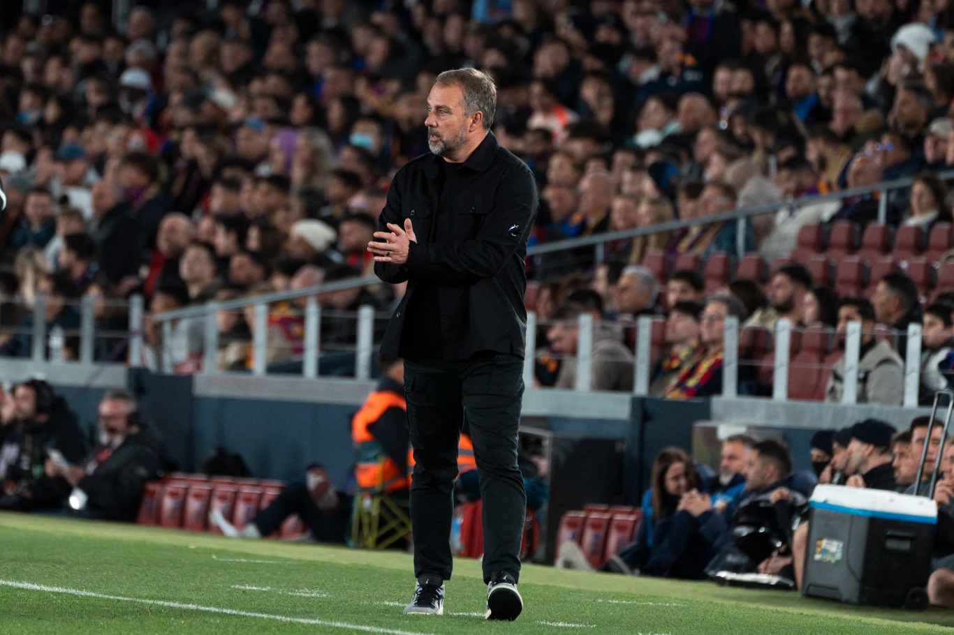 Trener drużyny piłkarskiej ubrany na czarno stoi przy linii bocznej boiska, klaszcząc w dłonie, w tle tłum kibiców na trybunach stadionu.