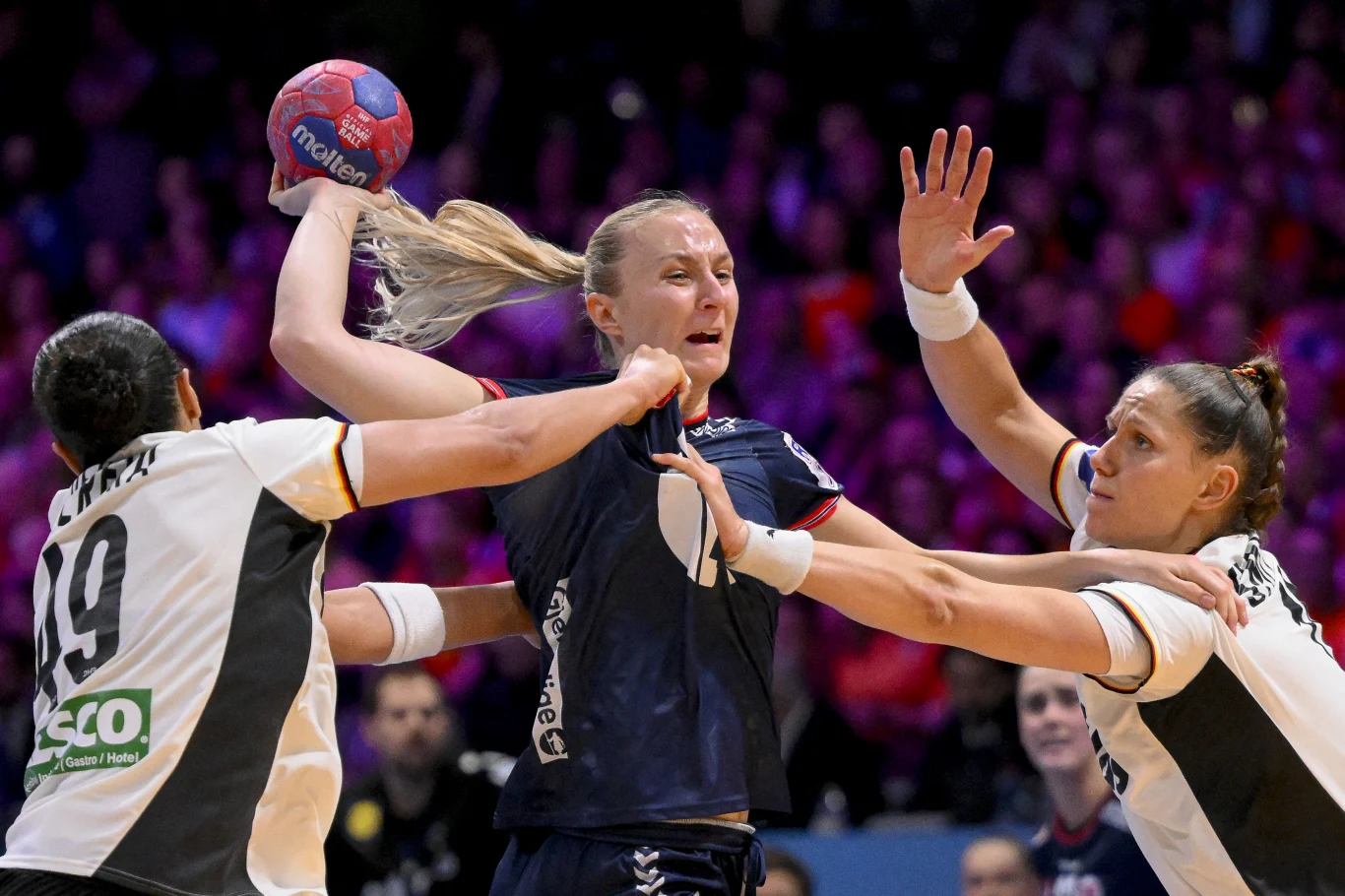 Dynamiczny moment podczas meczu piłki ręcznej z udziałem trzech zawodniczek, dwóch w białych koszulkach próbujących zablokować przeciwniczkę w ciemnoniebieskim stroju, która podnosi piłkę do rzutu.