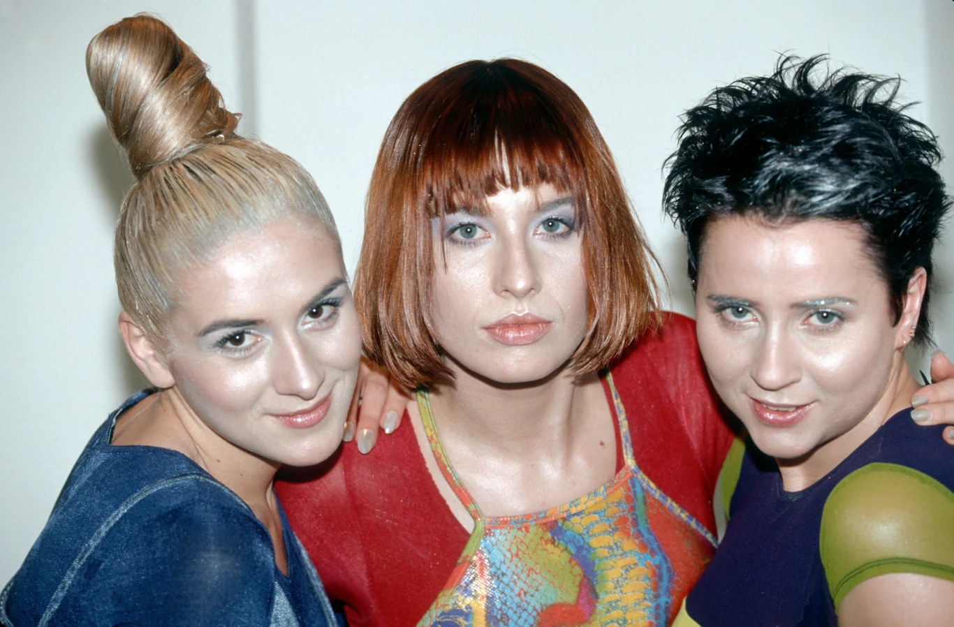 Zespół De Su - (od lewej) Daria Druzgała, Małgorzata Pruszyńska, Beata Kasprzyk Trzy kobiety pozujące razem, ubrane w kolorowe, nowoczesne stroje, zabawne fryzury w stylu lat 90-tych, jasne tło oraz przyjazne, uśmiechnięte wyrazy twarzy.