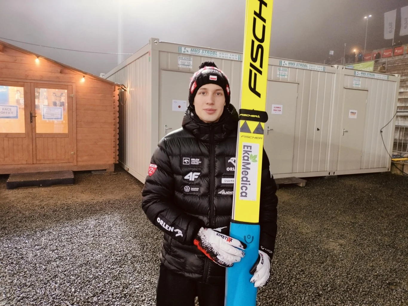 Młody skoczek narciarski w zimowej kurtce, rękawiczkach i czapce z logotypami sponsorów trzyma długie narty Fischer. W tle znajdują się kontenery techniczne oraz drewniany domek oświetlony lampą.
