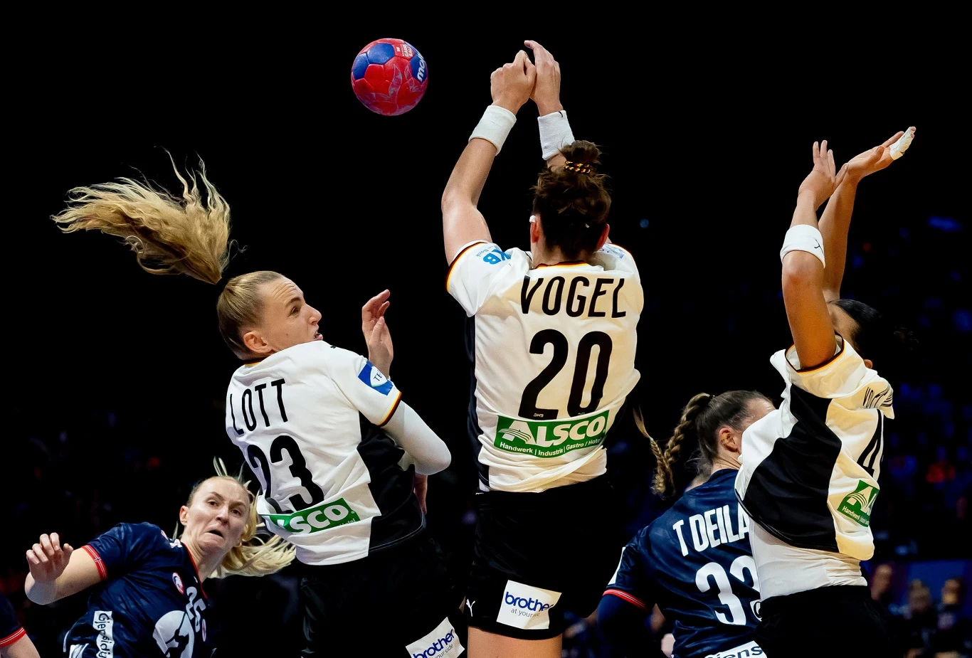 Zawodniczki dwóch drużyn piłki ręcznej walczące o piłkę w trakcie dynamicznej akcji podczas meczu, w centrum ujęcia trzy zawodniczki w białych koszulkach, widoczne wyskoki i zacięcie sportowe.
