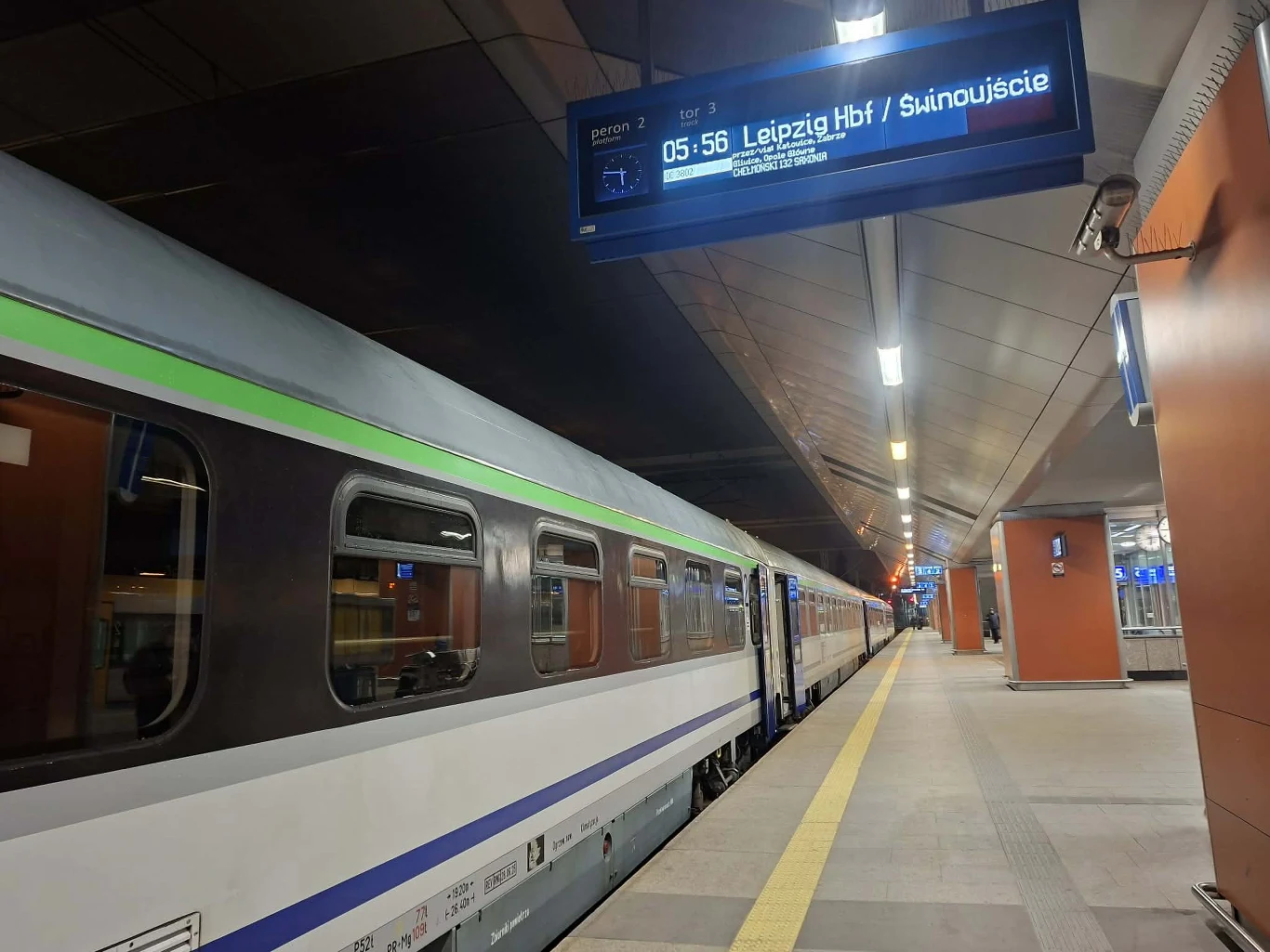 Pociąg pasażerski stojący na peronie w oświetlonej hali dworca, nad peronem cyfrowy wyświetlacz informacyjny z nazwami stacji Leipzig Hbf oraz Świnoujście, w tle widać nowoczesne elementy infrastruktury dworcowej.