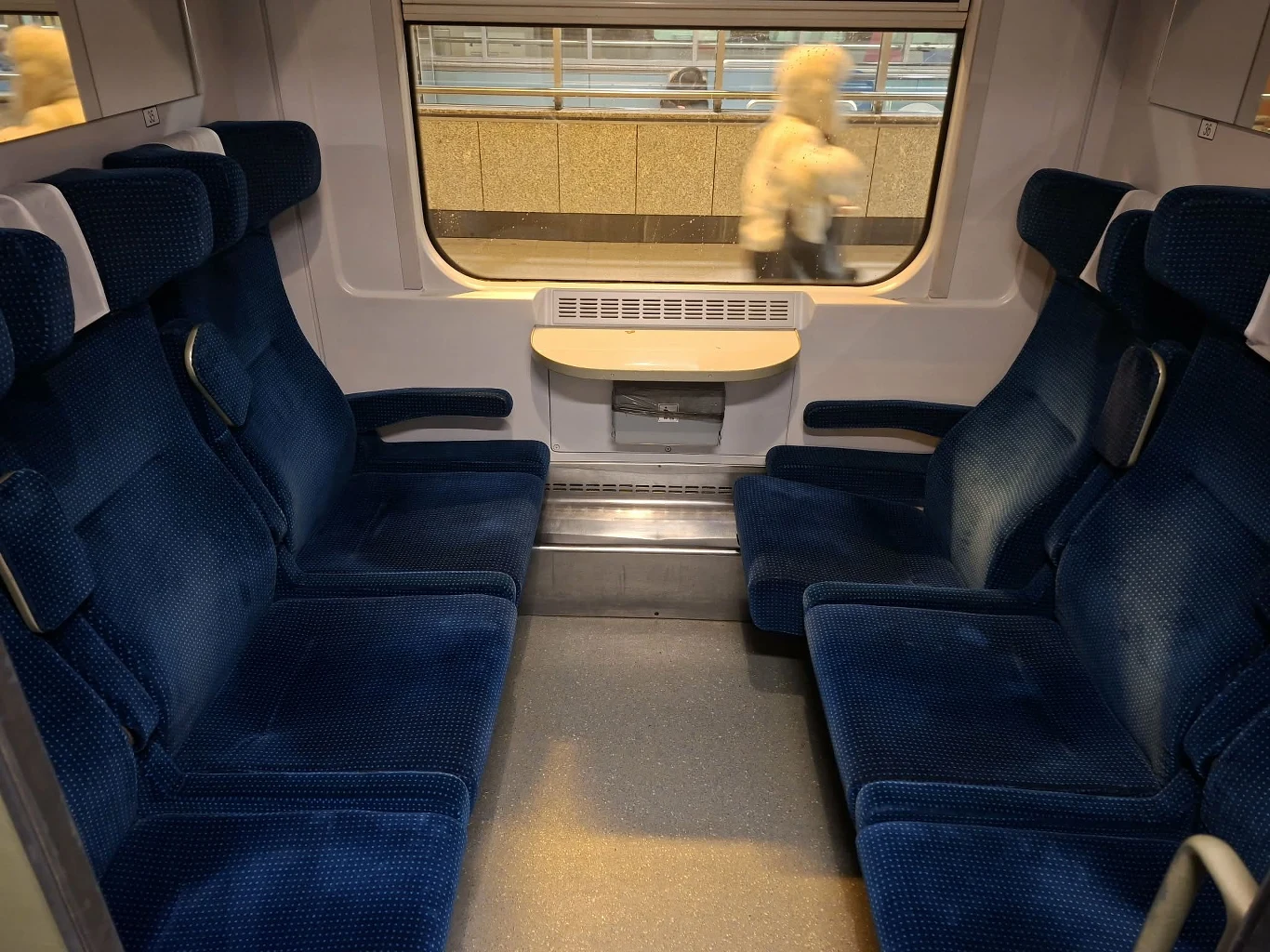 Wnętrze przedziału drugiej klasy w IC/EC "Saxonia" Przedział w nowoczesnym wagonie kolejowym z czterema miejscami po każdej stronie, składane podłokietniki, niebieskie tapicerowane siedzenia, stolik pod dużym oknem, za którym widoczny jest peron i sylwetka przechodnia.