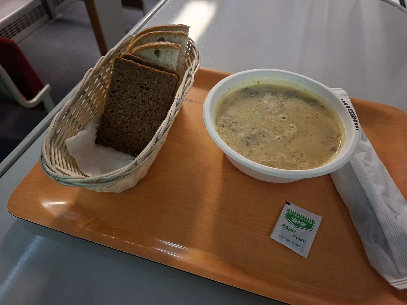 Smacznego :) Porcja zupy w jednorazowym pojemniku ustawiona na tacy obok koszyka z ciemnym pieczywem oraz saszetki soli i plastikowych sztućców.