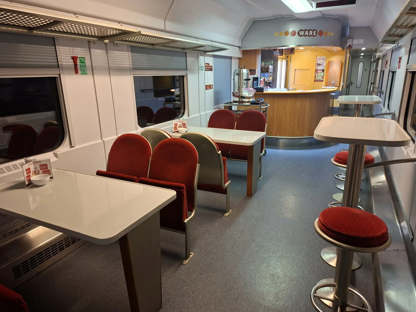 Wnętrze Warsu w IC "Chełmoński" / "Saxonia" Wnętrze wagonu restauracyjnego w pociągu z czerwonymi tapicerowanymi krzesłami, nowoczesnymi stołami oraz barem na końcu wagonu z wysokimi stołkami. Jasne oświetlenie i czysto utrzymana przestrzeń.