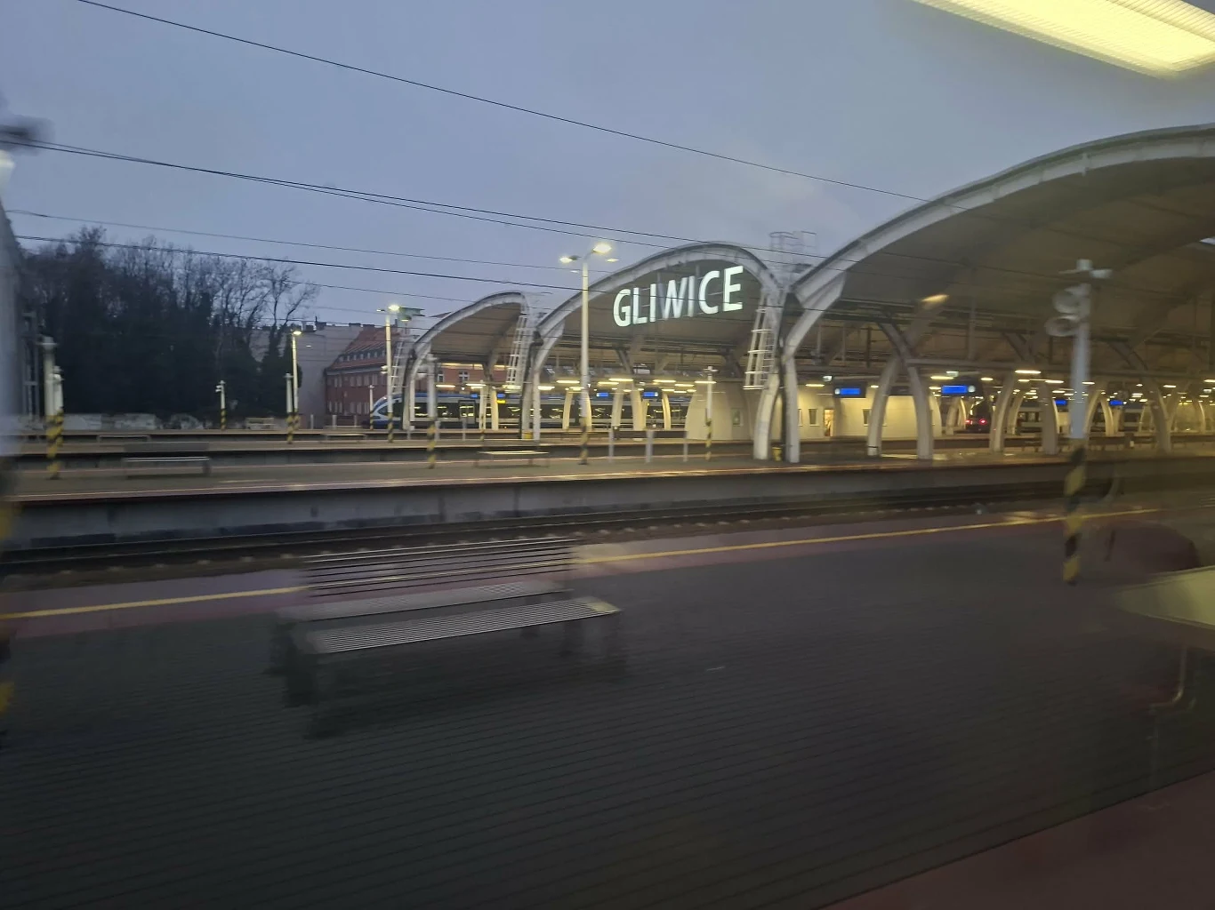 Jedną ze stacji odwiedzanych przez pociąg do Lipska są Gliwice Peron dworca kolejowego w Gliwicach podczas szarości dnia, z widocznymi ławkami, torami oraz zadaszeniem peronu i wyraźnie podświetlonym napisem Gliwice.