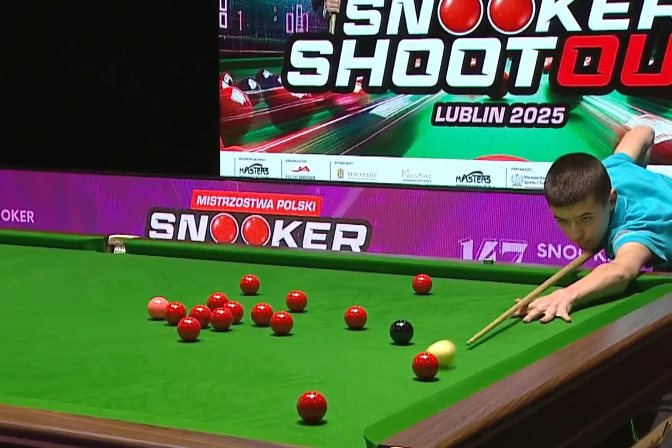 Zawodnik w niebieskiej koszulce wykonuje uderzenie kijem bilardowym podczas meczu snookera na profesjonalnym stole, w tle widoczna jest tablica z informacją o mistrzostwach i kolorowa grafika promująca wydarzenie.