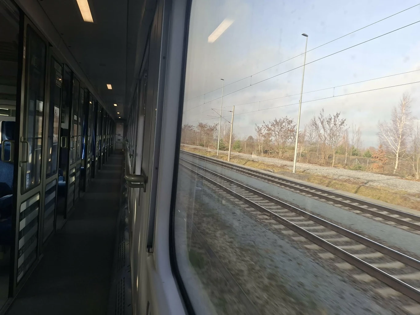 To już niemieckie tory Wnętrze nowoczesnego pociągu o pustych siedzeniach, widok przez okno na dwa tory kolejowe biegnące wzdłuż nasypu, w tle suche drzewa i słupy trakcji elektrycznej.